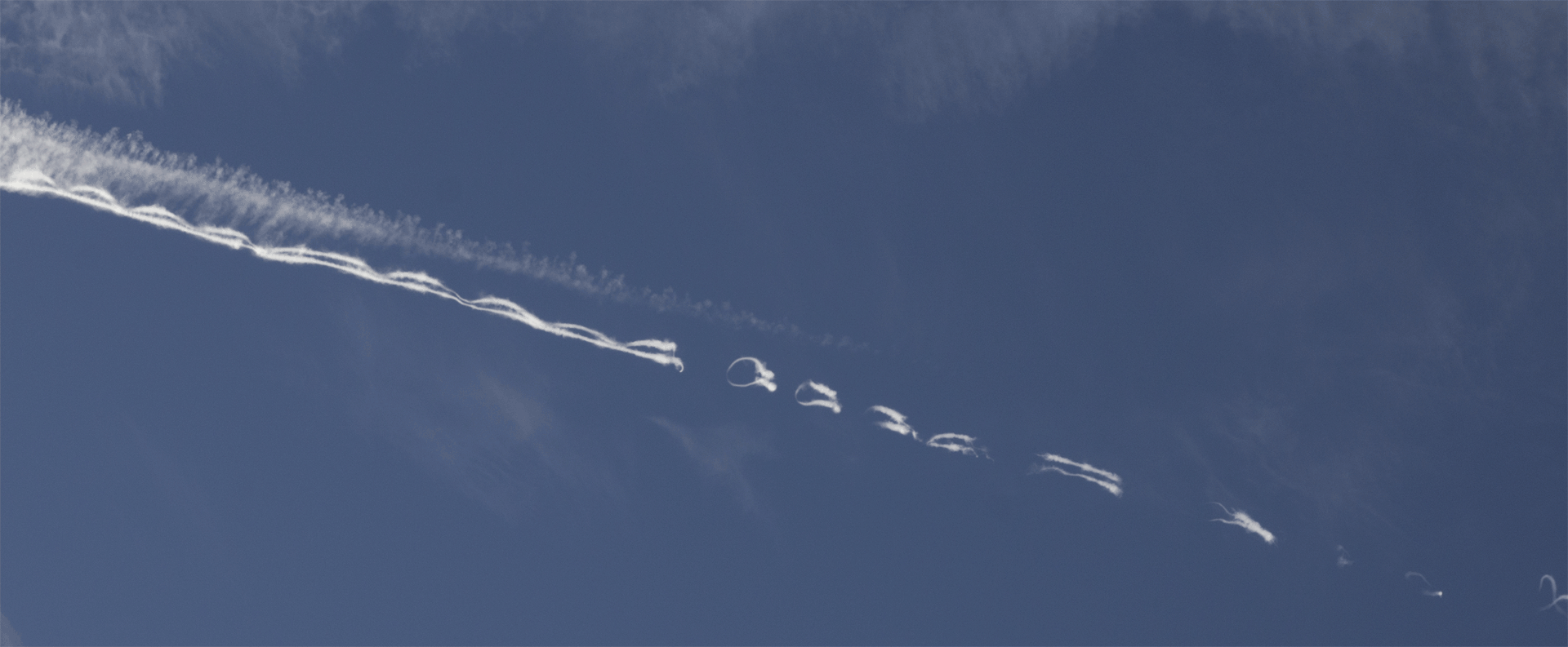 Persistent aircraft contrail exhibiting Crow instability as wingtip vortices deform the trail into repeating loops and wave-like structures at cruising altitude.
